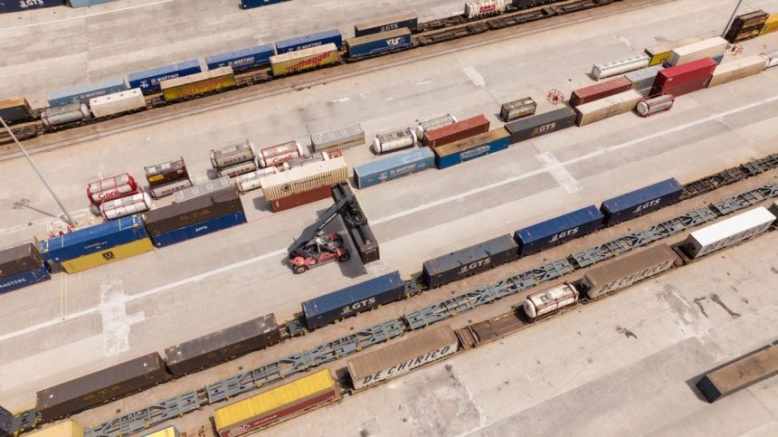 Vista aérea de um terminal logístico com contêineres empilhados e uma empilhadeira em operação.